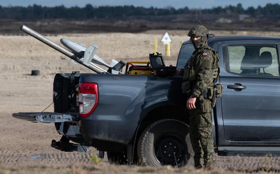 A soldier in camouflage uniform stands next to a dark-colored pickup truck in a grassy field with a military drone in the bed of the truck pointed toward the sky.
